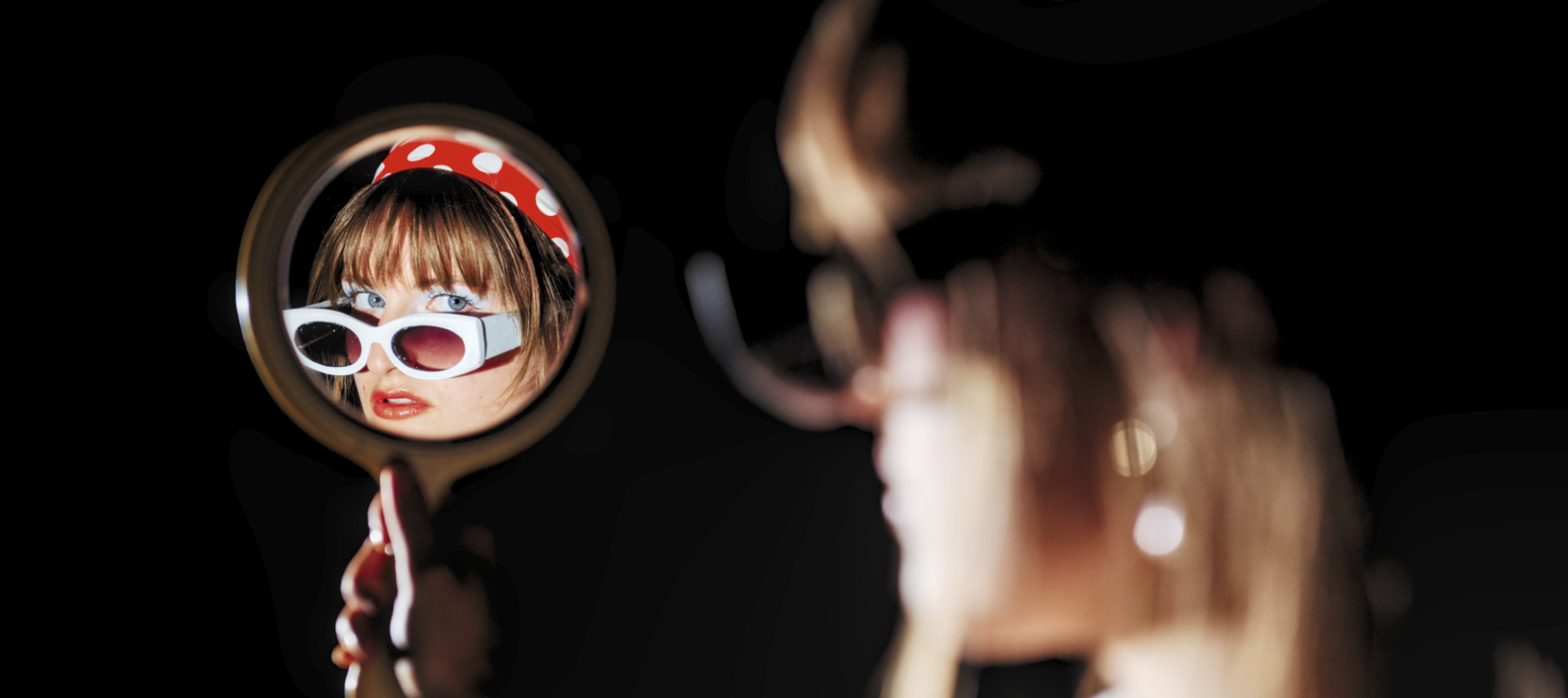 A student model poses in sunglasses and a red and white polka dot headband via a handheld mirror
